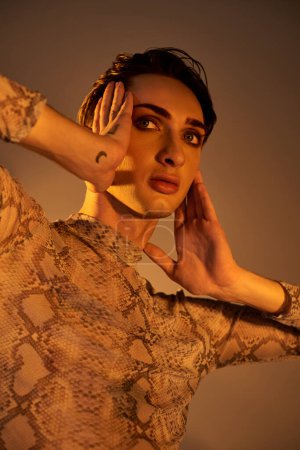 Eine junge Queer in einem Kleid aus Schlangenhaut posiert mit den Händen an den Ohren in einem stilvollen, stolz inspirierten Ensemble.