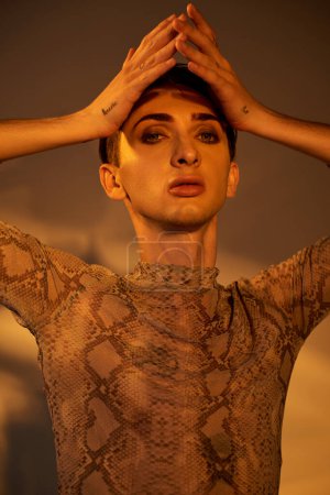 A young queer person in a snake skin top poses with hands on head in a fierce display of style and confidence.