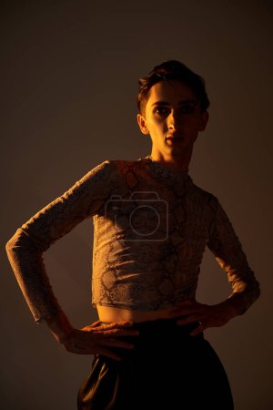 A young queer person strikes a stylish pose in front of a dark background, exuding confidence and pride.
