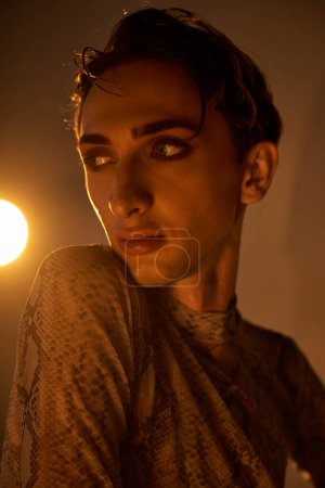 Un joven queer con un atuendo elegante se para con confianza frente a una luz radiante en una habitación tenue.