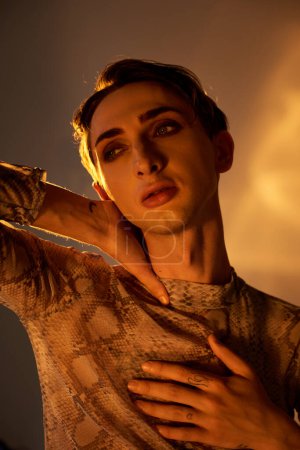 A young queer man in a snake skin shirt strikes a pose with hands on chest, showcasing pride and style.