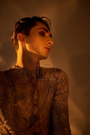 A young queer man in a snakeskin shirt stands confidently in a dimly lit room.