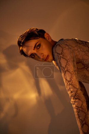 Eine junge queere Person in einem Kleid aus Schlangenhaut lehnt an einer Wand und strahlt Stil und Zuversicht aus.