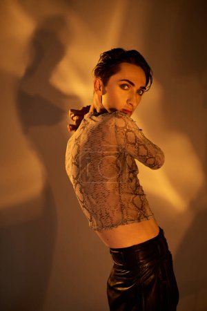 Eine stylische junge queere Person im Lederrock posiert selbstbewusst vor einer strukturierten Wand.
