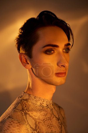 A young queer man in a snake skin dress stands gracefully in front of a bright light, exuding pride and style.