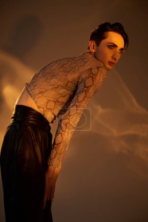 Una persona queer joven con confianza se encuentra en pantalones negros con estilo y una parte superior de piel de serpiente, exudando orgullo e individualidad.