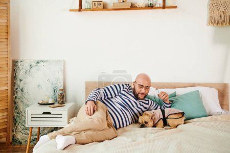Téléchargez les photos : Beau homme avec des lunettes couché sur un lit à côté d'un bouledogue français. - en image libre de droit