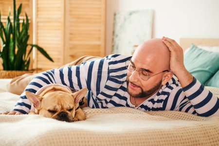 A man with glasses lounging on a bed alongside a French bulldog.