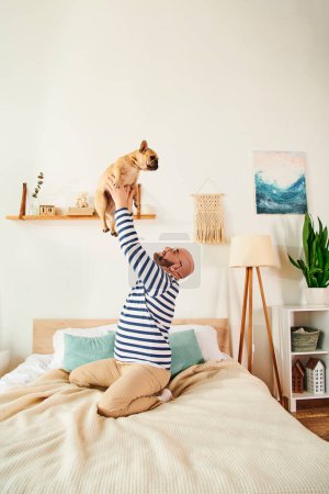 Foto de Hombre relajado con gafas levanta amorosamente Bulldog francés en el aire en la cama acogedora. - Imagen libre de derechos
