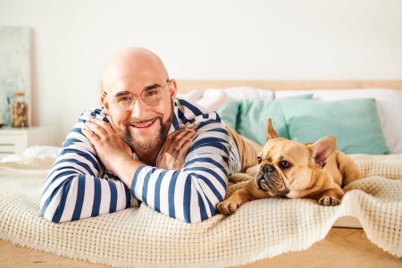 Schöner Mann mit Brille entspannt auf dem Bett neben seiner treuen französischen Bulldogge.