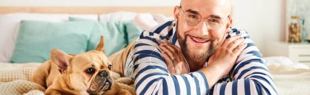 Foto de Un hombre en gafas se relaja junto a su bulldog francés en una cama. - Imagen libre de derechos