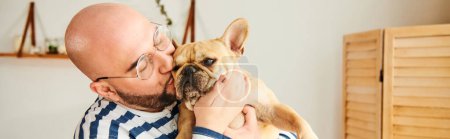 Un hombre con gafas sostiene un pequeño bulldog francés en sus brazos.