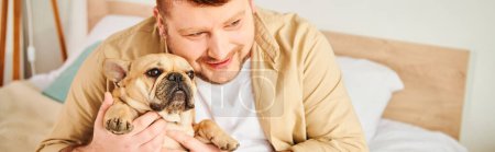 Handsome man tenderly holds a small French Bulldog in his arms at home.