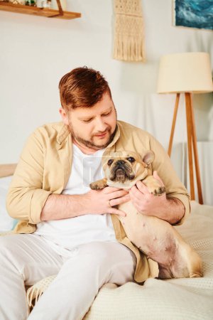 A man sitting on a bed, lovingly holding a small French bulldog.