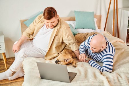 Two men and a dog relax on a bed with a laptop.