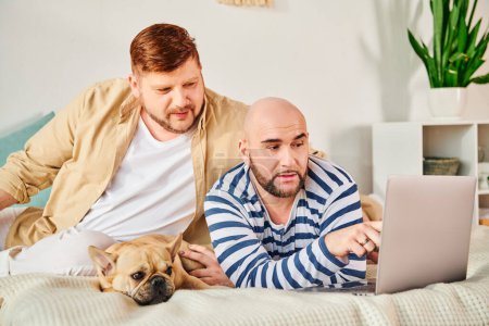 Two men and dog bond over laptop on bed.