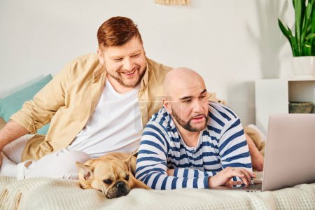 Two men and his dog are captivated by a laptop on a bed.