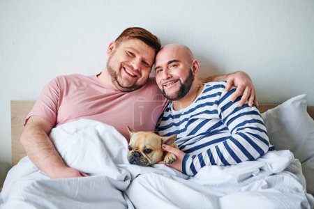 Two men cuddle in bed with their French bulldog.