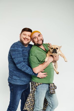 Foto de Dos hombres abrazan mientras sostienen un lindo Bulldog francés. - Imagen libre de derechos