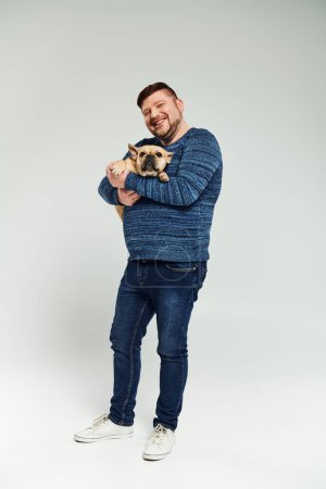A man tenderly holds a small French bulldog in his arms.