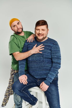 Two men posing together on a stool and looking at camera.