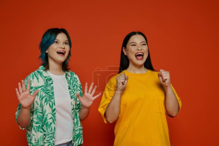 Two women of Asian descent stand with hands raised in a studio setting against an orange background.