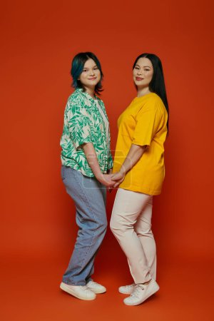 Foto de Una madre asiática y su hija adolescente de pie con gracia una al lado de la otra sobre un fondo rojo vibrante. - Imagen libre de derechos