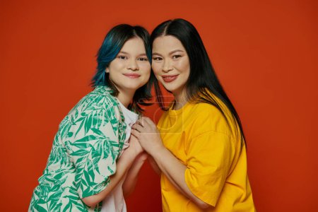 Foto de Una madre asiática y su hija adolescente se paran una al lado de la otra frente a una llamativa pared roja. - Imagen libre de derechos