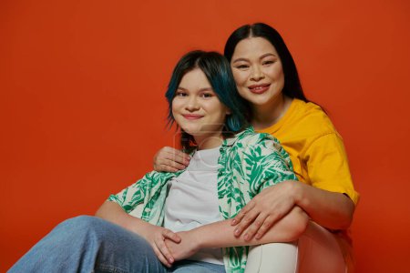 An Asian mother and her teenage daughter sit gracefully on a chair against a vibrant red background.