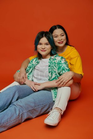 An Asian mother and her teenage daughter sit on the ground and pose for a picture against an orange studio background.