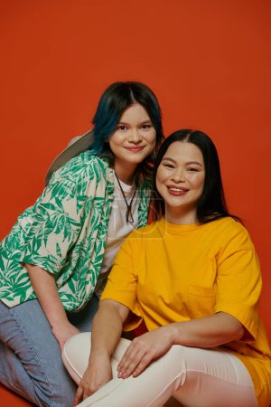 Foto de Una madre asiática y su hija adolescente sonríen, sentadas una al lado de la otra sobre un fondo naranja, posando juntas para una foto. - Imagen libre de derechos