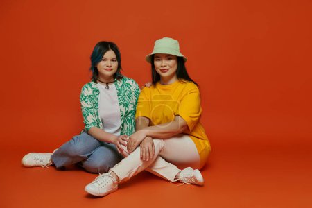 eine asiatische Mutter und ihre Teenager-Tochter, die auf dem Boden sitzt und anmutig in einem Studio auf orangefarbenem Hintergrund posiert.