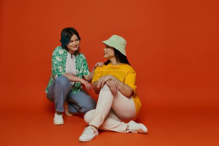Asiatische Mutter und ihre Teenager-Tochter sitzen eng beieinander und teilen einen Moment der Verbundenheit auf einem leuchtend orangefarbenen Hintergrund.