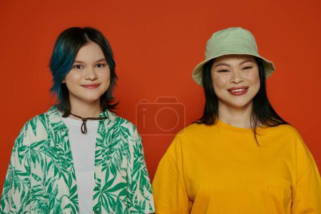 Photo for An Asian mother and her teenage daughter standing side by side in front of a vibrant red wall. - Royalty Free Image