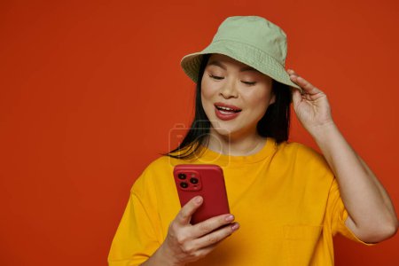 Foto de Una mujer asiática de moda en un sombrero sosteniendo un teléfono celular mientras está sobre un fondo naranja. - Imagen libre de derechos