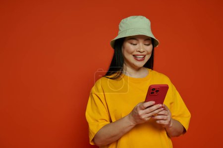 Asian woman wearing a yellow t-shirt joyfully holds a red cell phone in a studio with an orange background.