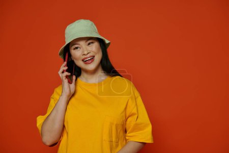 A woman in a yellow shirt speaks on a cell phone on an orange background.
