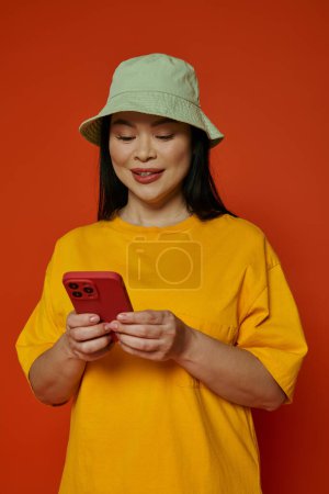 A woman in a hat browsing her cell phone in a studio with an orange backdrop.