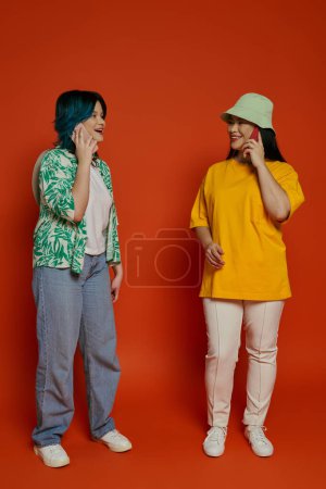 a mother and her teenage daughter, standing together on an orange background, engaged in separate phone conversations.