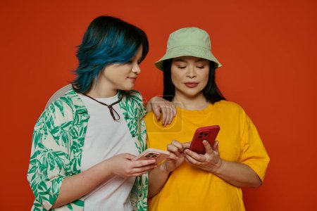 Téléchargez les photos : Mère asiatique et sa fille adolescente debout ensemble, regardant un écran de téléphone portable, sur un fond orange. - en image libre de droit