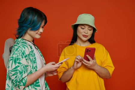 Asiatische Mutter und ihre halbwüchsige Tochter stehen zusammen, in einen Handy-Bildschirm vertieft, auf orangefarbenem Studiohintergrund.