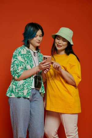 Foto de Una madre asiática y su hija adolescente de pie juntas, absortas mientras miran un teléfono celular sobre un fondo naranja. - Imagen libre de derechos