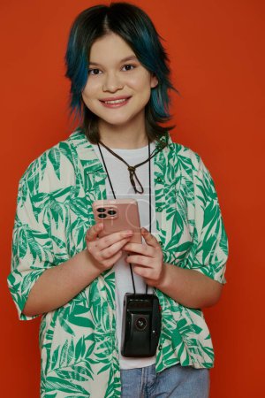 Téléchargez les photos : Une adolescente asiatique créative aux cheveux bleus tenant un téléphone portable dans un studio sur fond orange. - en image libre de droit