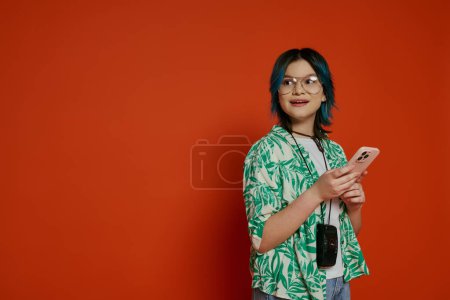 Foto de Una chica adolescente con estilo con el pelo azul y gafas sostiene un teléfono celular en un entorno de estudio vibrante. - Imagen libre de derechos