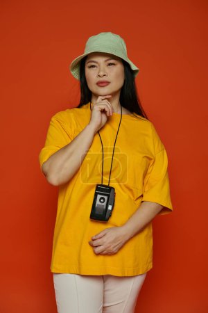 Foto de Mujer elegantemente vestida con una camisa amarilla y un sombrero verde sobre un fondo naranja. - Imagen libre de derechos