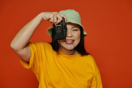 An Asian woman taking a picture, using a retro camera on an orange background.