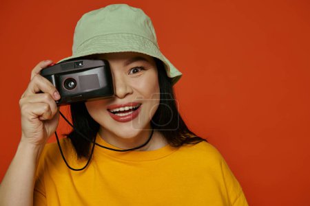 A woman holding a camera up to her face in a studio on an orange background.