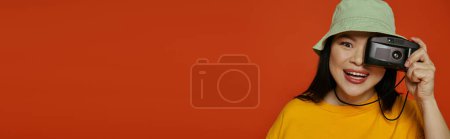 An Asian woman holds a camera to her face in a studio against an orange backdrop.
