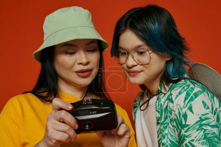 Foto de Dos mujeres, una madre asiática y su hija adolescente, absortas en mirar una cámara de teléfono celular juntas sobre un fondo naranja. - Imagen libre de derechos