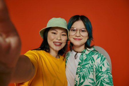 An Asian mother and her teenage daughter taking a selfie in a studio against an orange background.